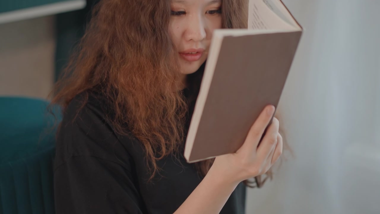 Close medium shot of woman seated indoors holding open book near face, eyes fixed on pages while hands grip cover, showing calm concentration in soft natural light and cozy home setting