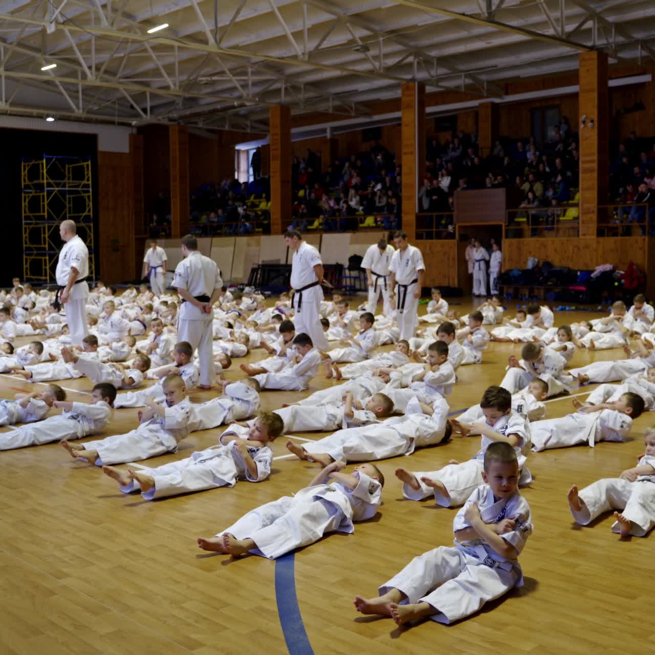 Large group of little athletes doing crunches lying on the wooden floor in gym. Team of coaches watch the trainees. Audience at backdrop