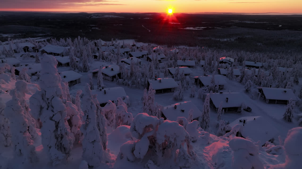 Ilmakuva lumisten puiden ympäriltä, paljastaen tunturimökkejä, talvinen auringonlasku Lapissa