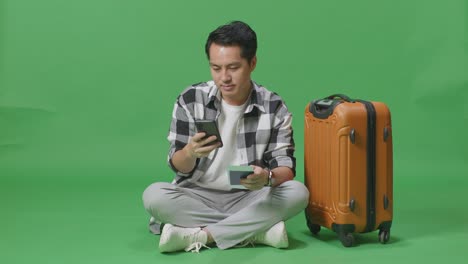 full body of asian male traveler with luggage holding passport and using smartphone while sitting in the green screen background studio