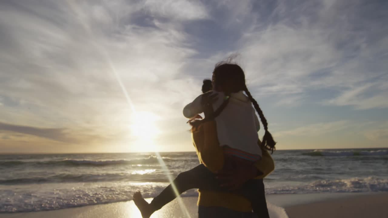 Back view of hispanic mother carrying piggyback daughter and looking at sunset on beach