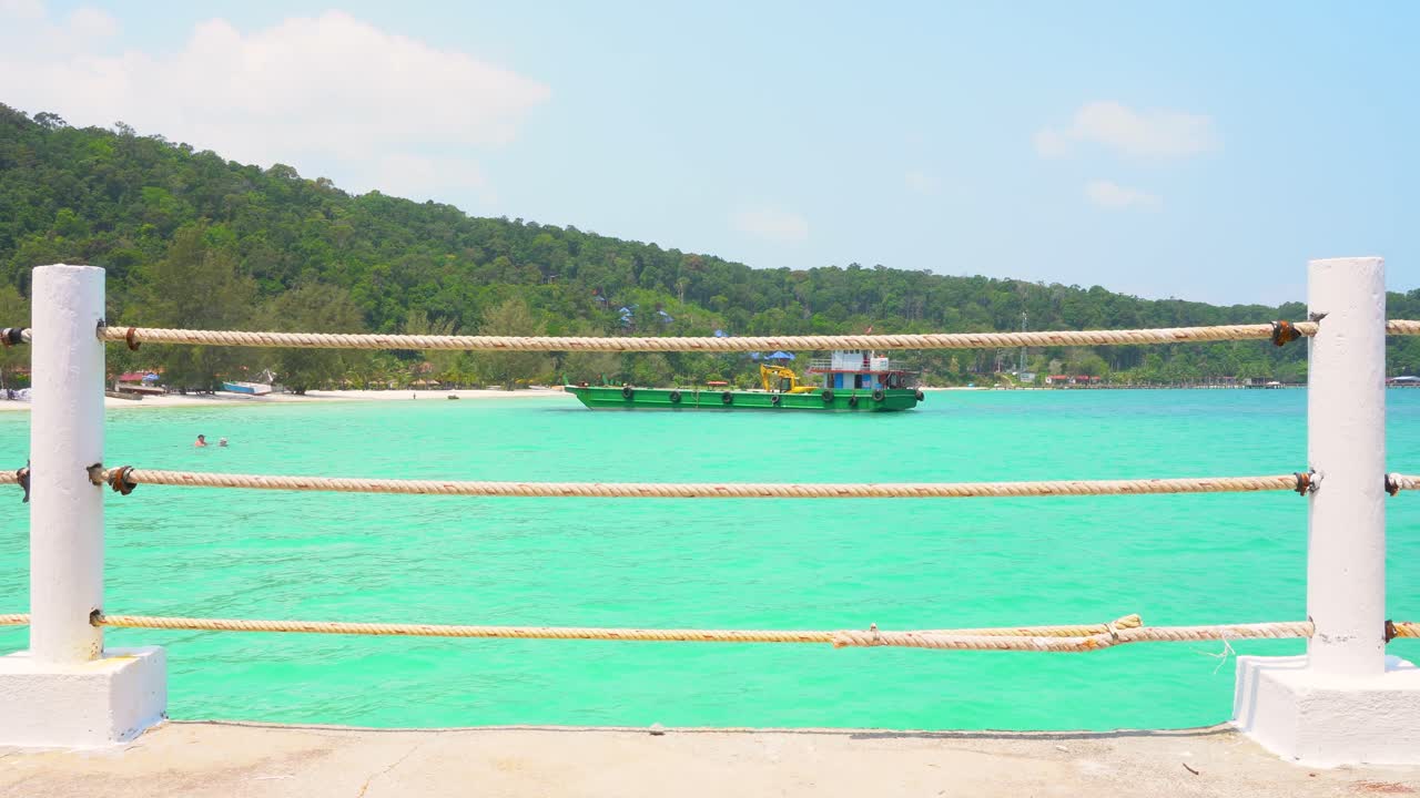 아름다운 캄보디아 해변에 정박한 건설 바지선은 아름다운 녹색 바다에 앉아 인근 부두의 로프 난간을 통해 볼 수 있습니다.