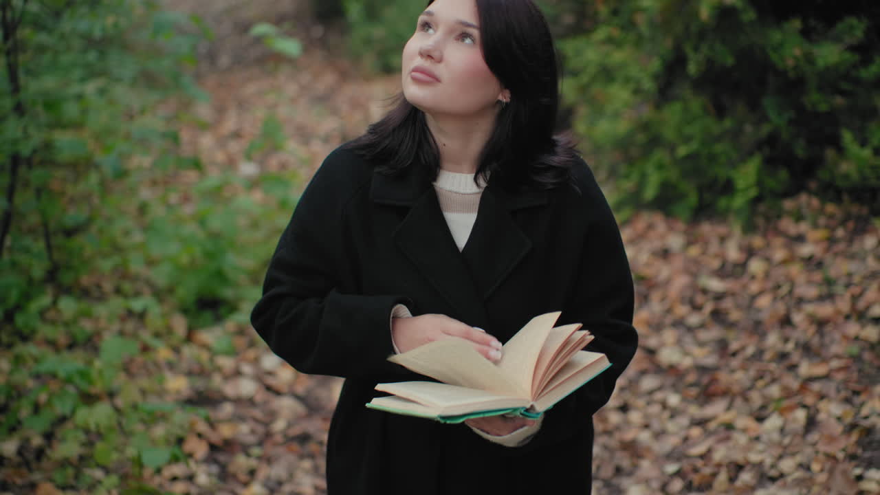 Vista elevada de una joven elegante con abrigo de otoño hojeando las páginas de un libro mientras pasea por un sendero forestal, ambiente tranquilo, mirada lenta alrededor, vegetación bokeh suave, lectura consciente, momento tranquilo al aire libre