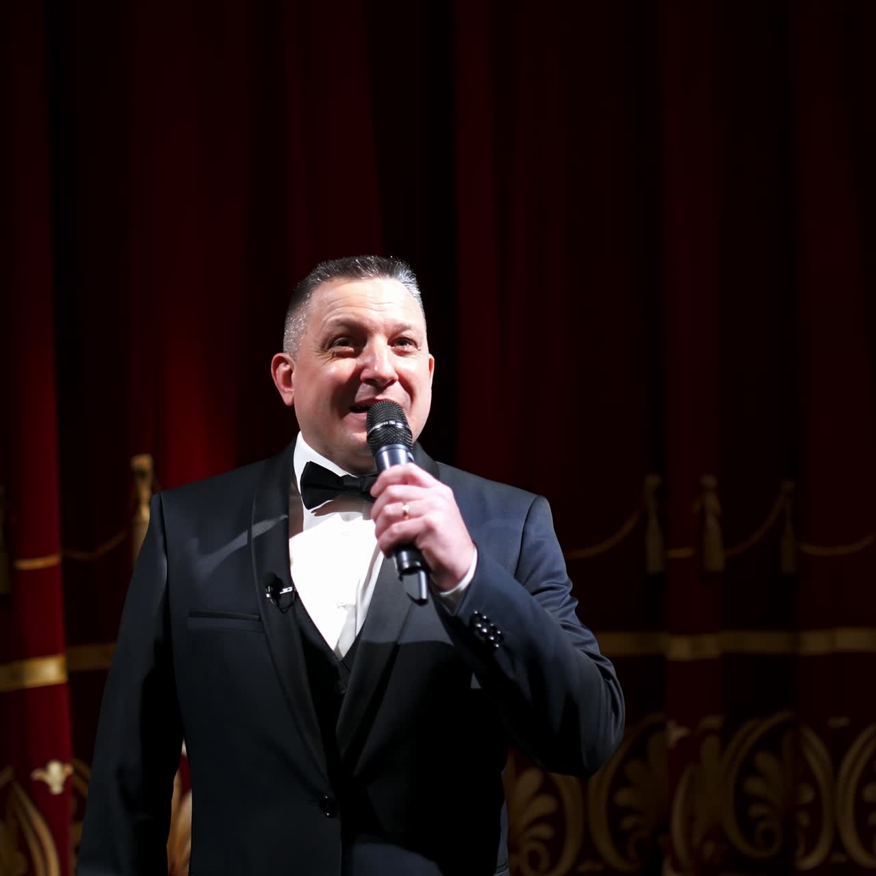 Host in elegant suit starts his speech. Speaker talking to the microphone on stage of a theater. Showman on red curtains background