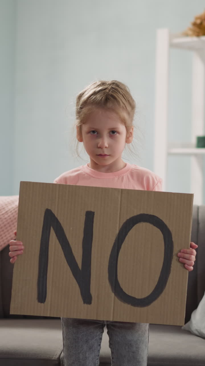 Offended little girl shows cardboard paper with word NO standing at home. Blonde child expresses protest against social events. Children protection
