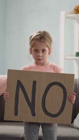 Offended little girl shows cardboard paper with word NO standing at home. Blonde child expresses protest against social events. Children protection