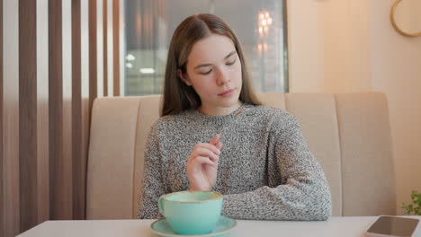 college graduate seated in restaurant holding spoon, carefully examining it with amazement, gently blowing it and returning it to tray on table while cars faintly move outside through glass