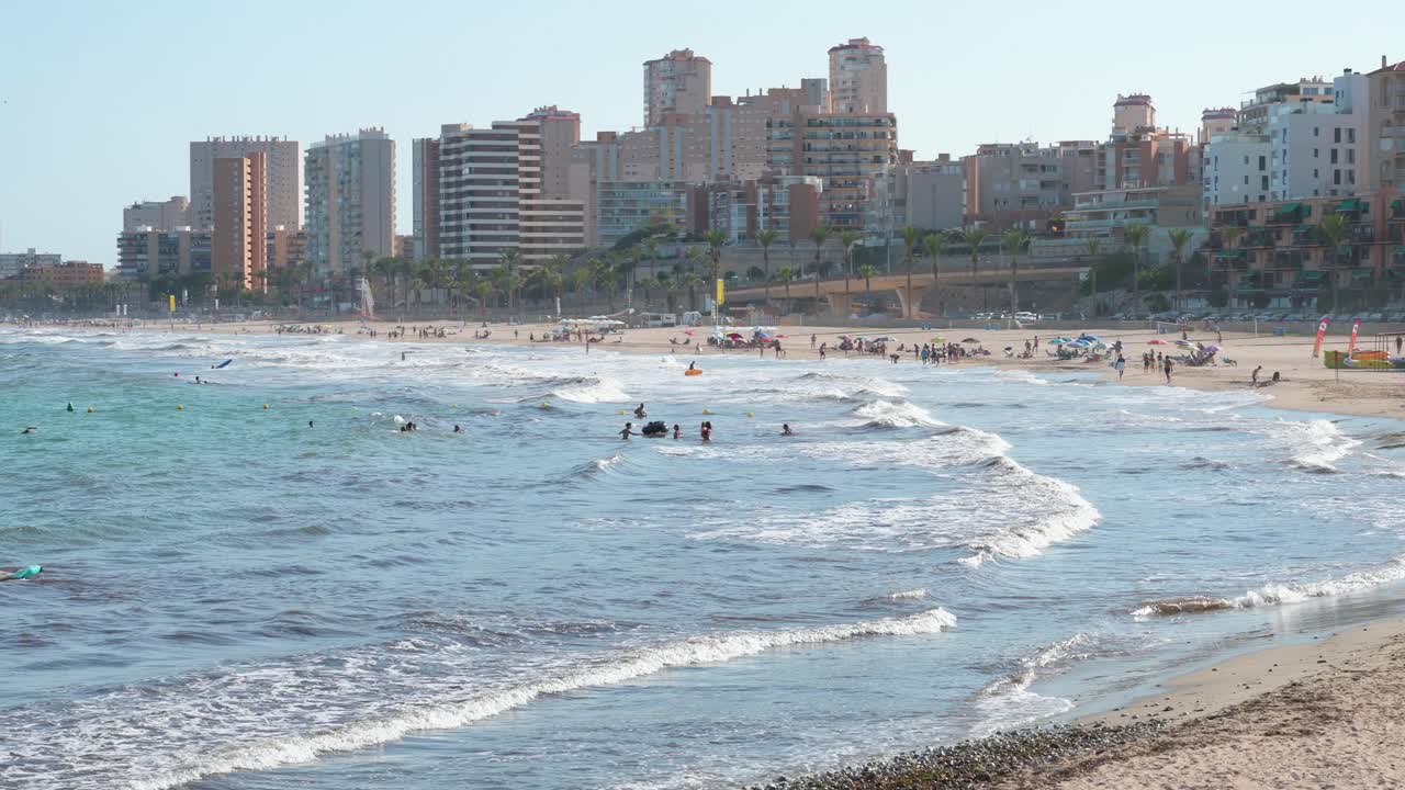 в эль-кампелло, коста-бланка, аликанте, средиземное море оживлено солнцезащитниками, наслаждающимися волнами и расслабляющимися на песке, в то время как пловцы веселятся рядом с берегом в испании.