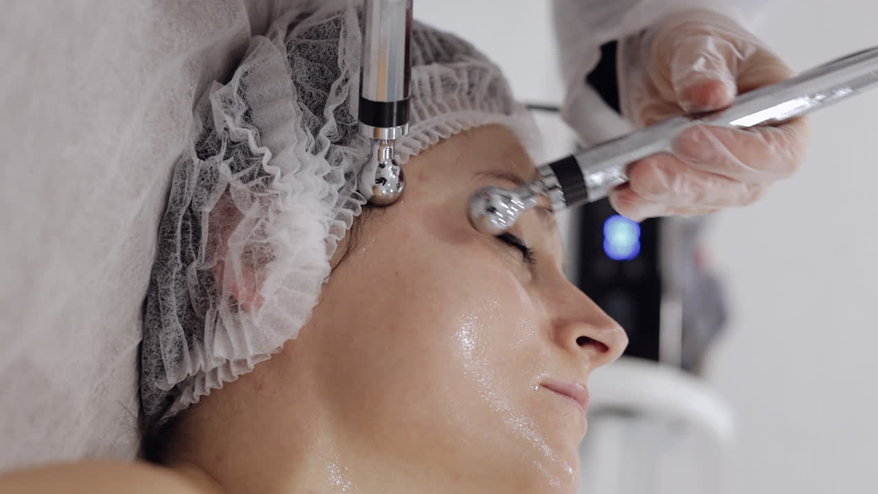 미용학자 의사는 스파 살롱에서 전기 충동을 사용하여 여성 마이크로 전류 얼굴 치료를합니다.