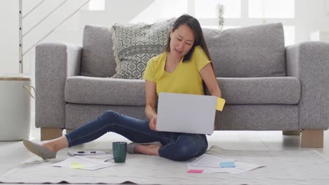 Mujer-Asiática-Enfocada-Sentada-En-El-Suelo-Y-Trabajando-Remotamente-Desde-Casa-Con-Una-Computadora-Portátil