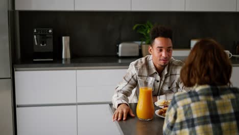 Over-the-shoulder-a-happy-young-guy-with-Black-skin-color-a-brunette-in-a-checkered-beige-shirt-communicates-with-his-girlfriend-with-brown-hair-and-a-bob-hairstyle-during-breakfast-in-a-modern-kitchen