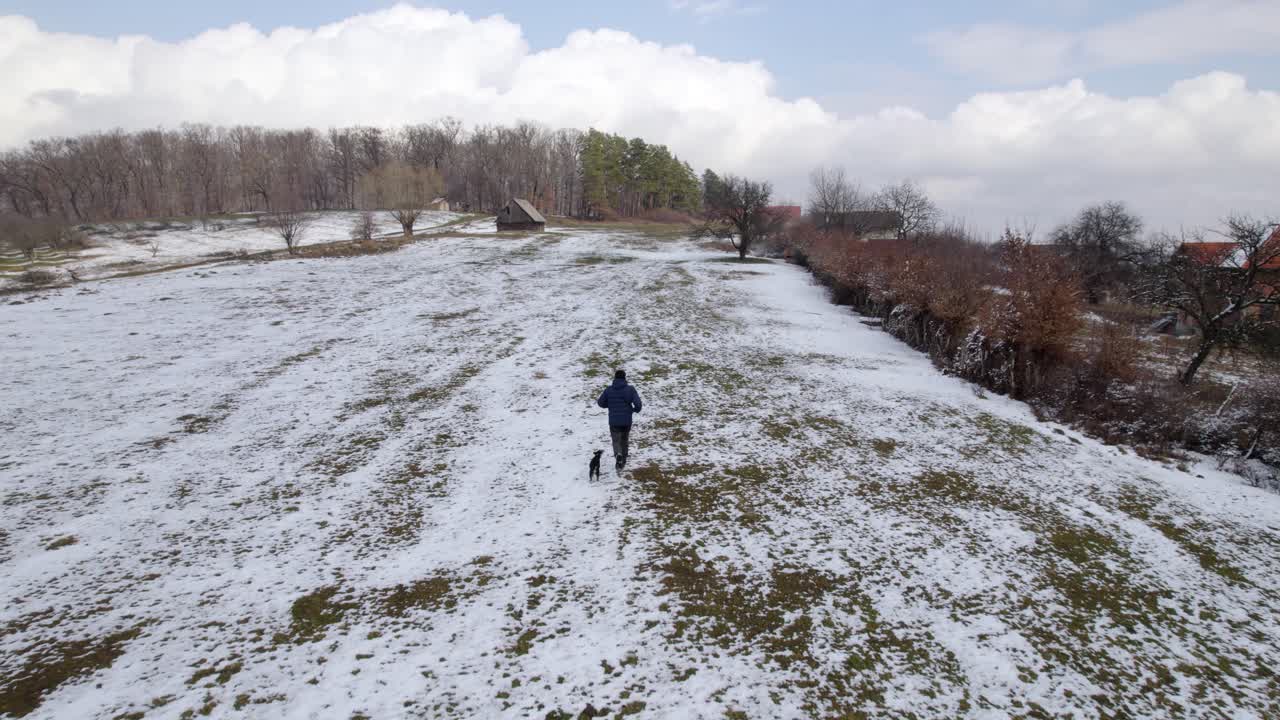 una persona y un pequeño cachorro corren a través de un paisaje nevado hacia una pequeña cabaña en invierno