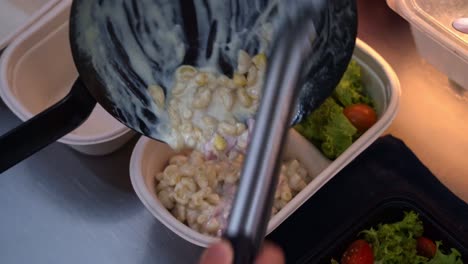 Chef-pouring-delicious-macaroni-and-cheese-into-an-eco-friendly-takeaway-container-with-salad-on-the-side,-restaurant-preparing-for-food-delivery