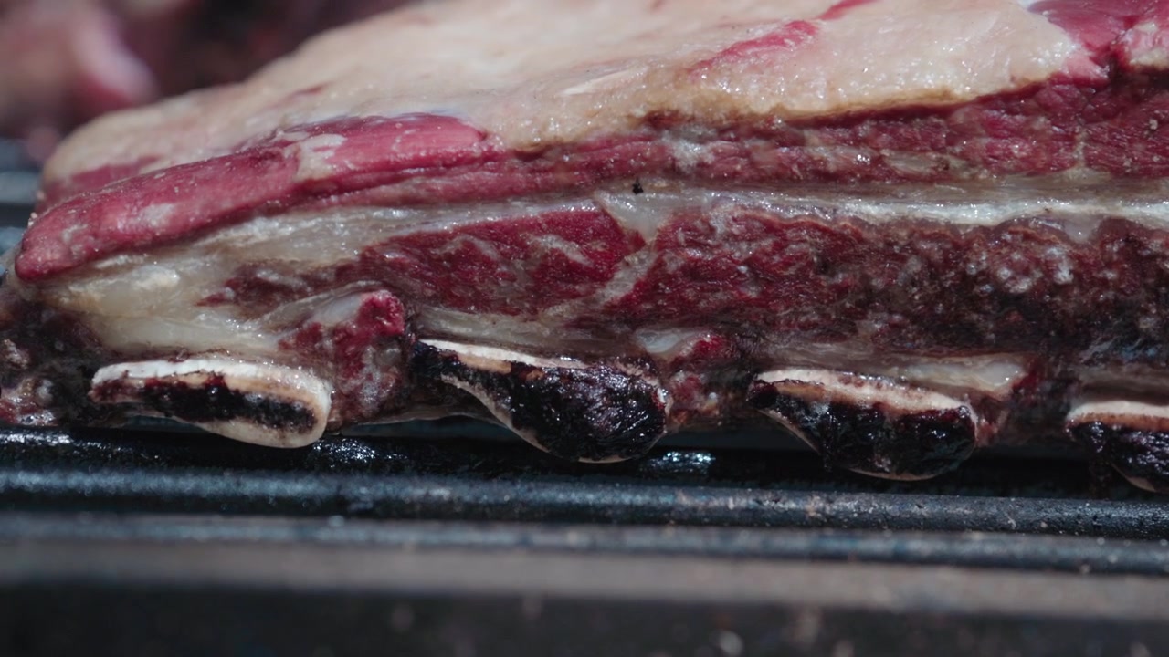Primo piano di asado de tira argentino che cuoce su una griglia rovente con grasso che sfrigola, fumo e calore intenso. Scena di barbecue rustico che mostra la cottura tradizionale a legna e la consistenza succosa della carne di manzo