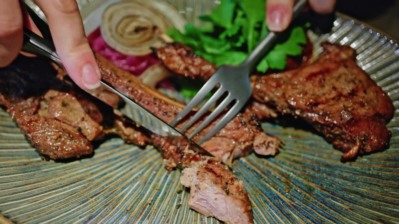 Gambar close-up iga babi panggang yang dilumuri bumbu gurih, sedang dipotong dengan pisau dan garpu di atas piring keramik