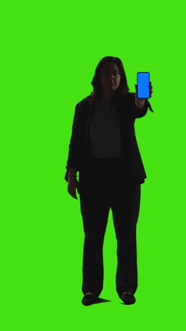 vertical video full length shot of businesswoman holding blue screen mobile phone towards camera standing against green screen