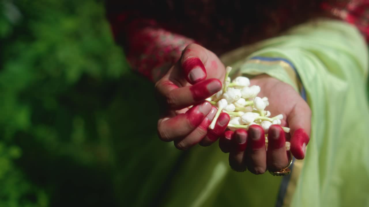Close-up of a woman wearing green and red traditional clothing, with henna-stained hands and gold jewelry, holding a handful of white jasmine buds, symbolizing purity, devotion and cultural tradition