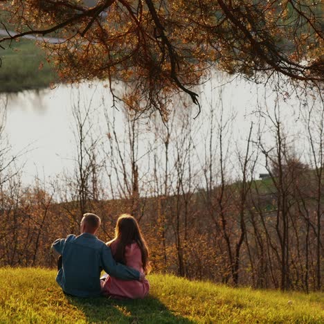 romantic couple enjoys the nature. young man and woman sitting on grass on the hill and looking at beautiful river in springtime. back view