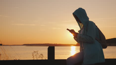 una mujer en una capucha se sienta en una cerca cerca de un lago pintoresco usa un hermoso atardecer de teléfono inteligente