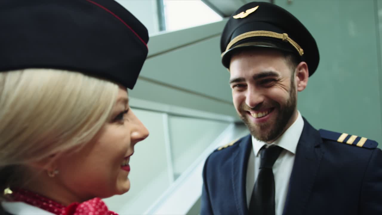 Na letišti se přívětivě usmívá letuška s blond vlasy a mluví s pilotem. Evropská žena oblečená v tmavě modré uniformě, šarlatově červený šátek uvázaný na bílé košili. Filmový záběr na kameru RED. 2 modelky