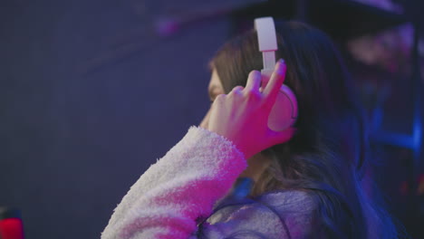 side profile of woman seated at tech desk placing white earphones while resting hand on desk under neon pink purple lighting preparing gaming setup for live stream broadcast in modern studio