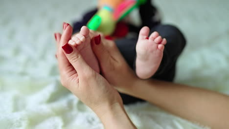 caring and loving mother's hands caressing bare feet of her son. newborn kid is busy with a toy while mum is rubbing tiny baby toes.