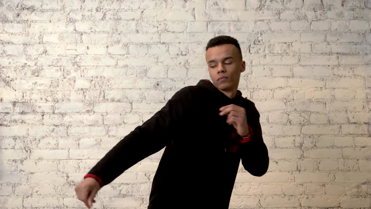 Giovane ragazzo africano con una felpa nera che balla impetuosamente contro un muro bianco. Concetto di discoteca, ballo, movimento, energia, gioventù. 60 fps