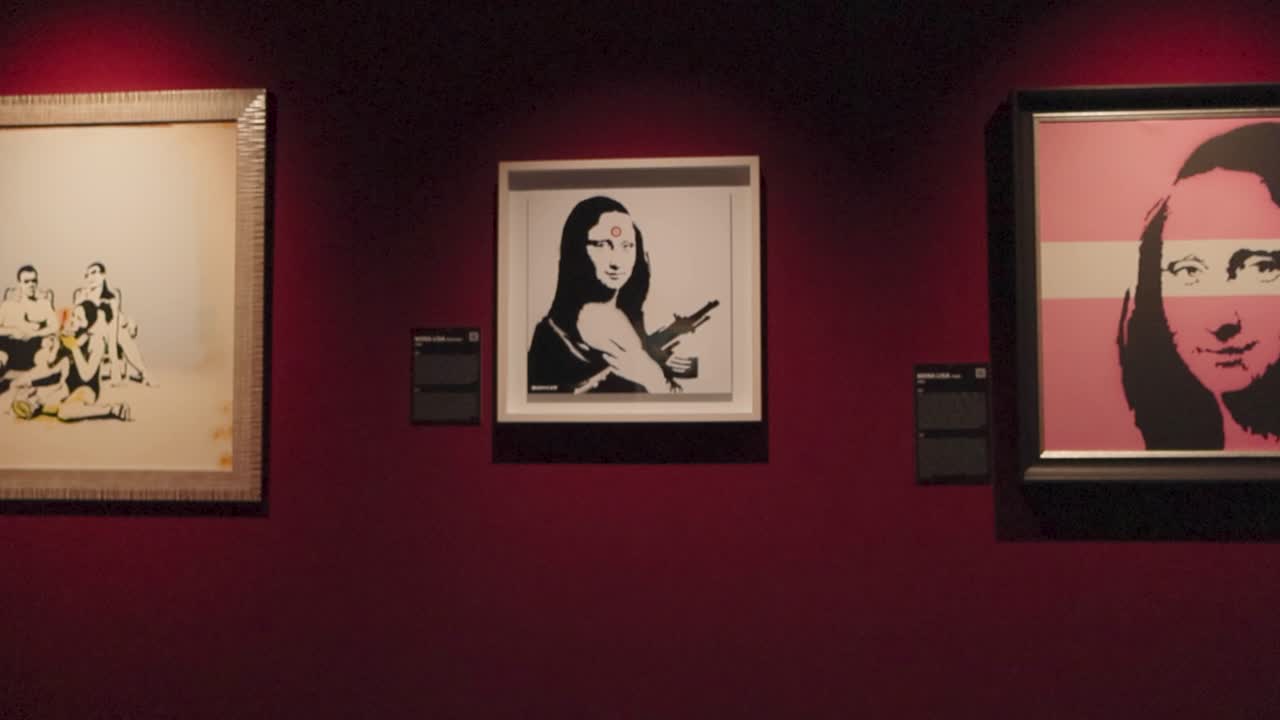 Red wall that has three Banksy framed spray paintings or graffiti artwork on it at his exhibition. Wall is red and two of the paintings show Mona Lisa slightly altered. descriptions are on the side.