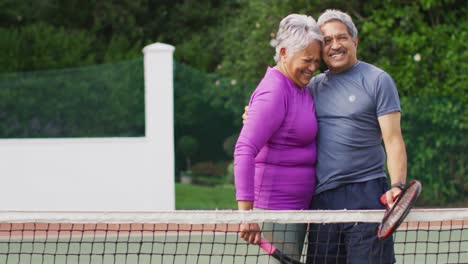 Video-Eines-Glücklichen-Gemischtrassigen-älteren-Paares,-Das-Sich-Während-Des-Trainings-Auf-Dem-Tennisplatz-Umarmt