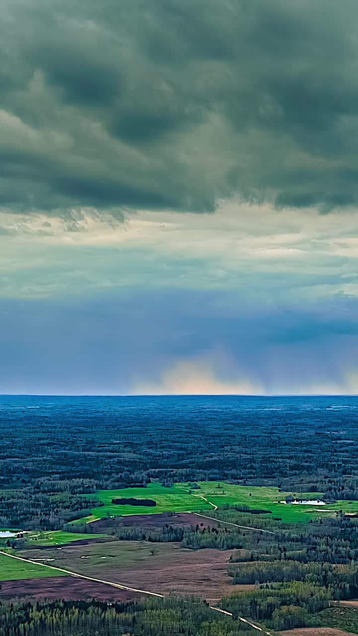 An aerial timelapse captures dark, dramatic storm clouds gathering and moving ominously across the sky above a vast forest and green fields in the remote Latvian countryside.