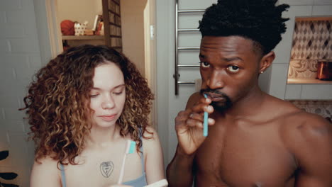 una pareja joven y diversa se cepilla los dientes y usa los teléfonos en el baño.