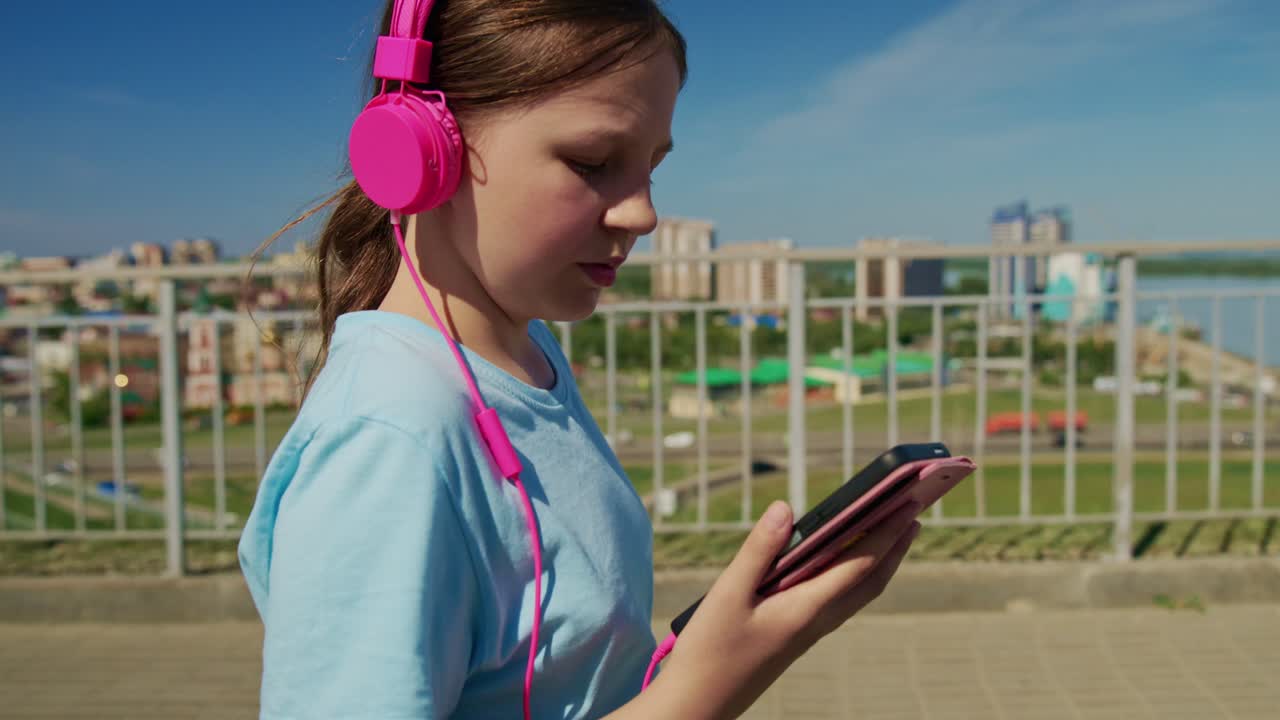 Una ragazza ascolta musica con cuffie rosa. Cammina vicino all'acqua con il telefono in mano. Colori vivaci e cielo aperto esaltano l'atmosfera. Ideale per contenuti di fitness, viaggi o lifestyle.