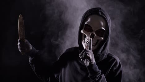 close up of scary man in the hooded sweatshirt wearing halloween mask gesturing silence while holding a knife on the black background with smoke