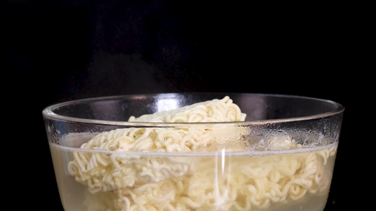 A block of instant noodles gradually softens and expands in a clear glass bowl of hot water, captured against a black background with steady lighting