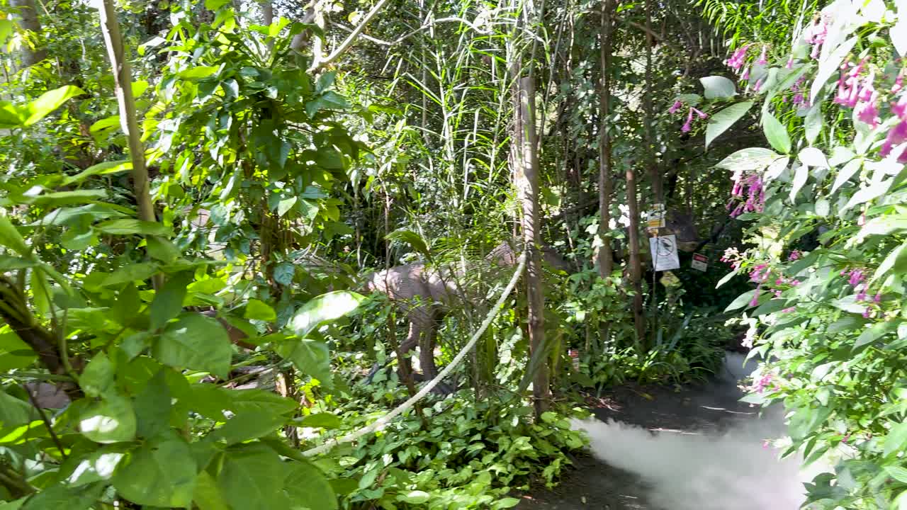 Camera moves through dense greenery, passing dinosaur models and vintage vehicle in bright daylight