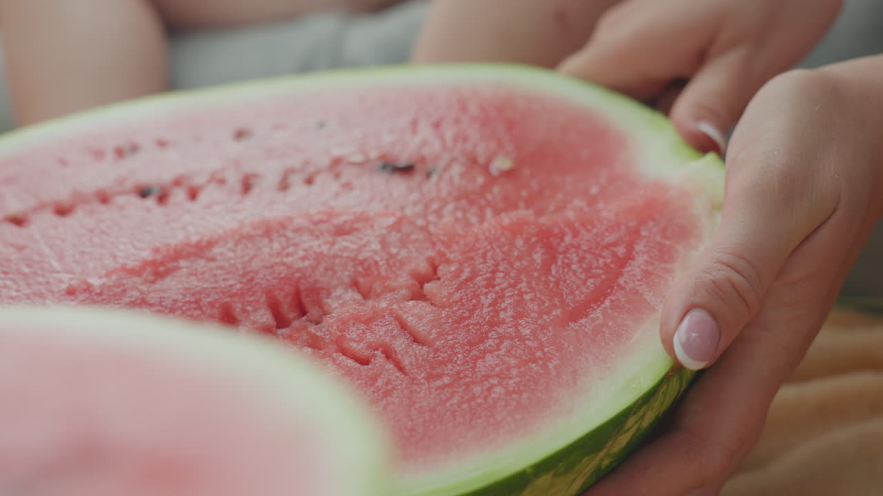 Caucasian hands slicing ripe watermelon closeup, knife gliding through red flesh on picnic blanket, focused prep and serving, careful grip, juice droplets, family gathering mood, outdoor sunlight