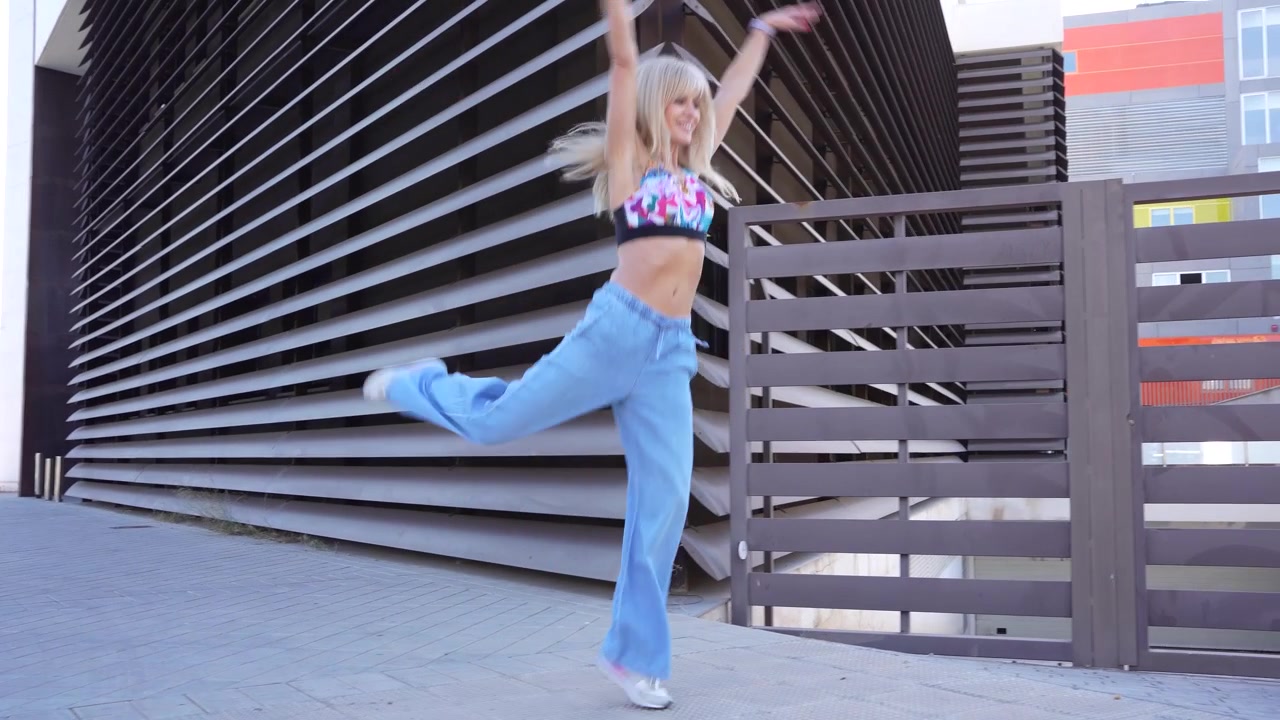 En blond kvinde danser glad i et bymiljø iført en crop top og jeans.