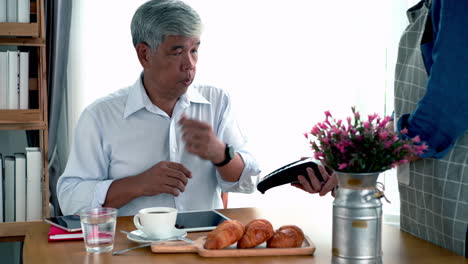 active senior asian man working in a coffeeshop using a mobile device, smartwatch for nfc, qr code scanning, contactless payment to pay for coffee and snack