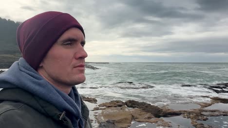 a caucasian male thoughtfully looks out at the sea as waves crash on the beach