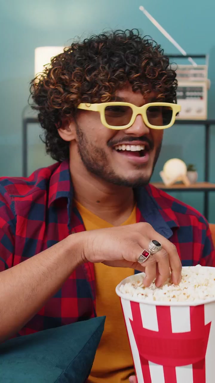 Indian man sits on couch eating popcorn and watching interesting tv serial sport game online at home