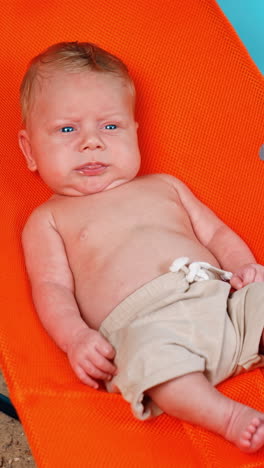 blond infant with blue eyes sits in the chair. infant in shorts resting on the improvised beach. vertical video