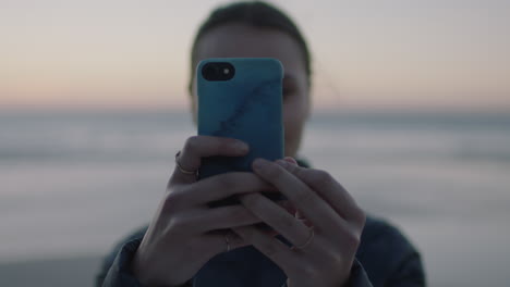 close up portrait of young woman taking photo using smartphone mobile camera technology on calm seaside beach at sunset