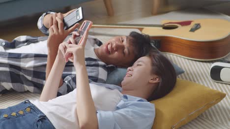 asian teen couple playing smartphone while lying on carpet on the floor at home. laughing, speaking, and enjoying time together