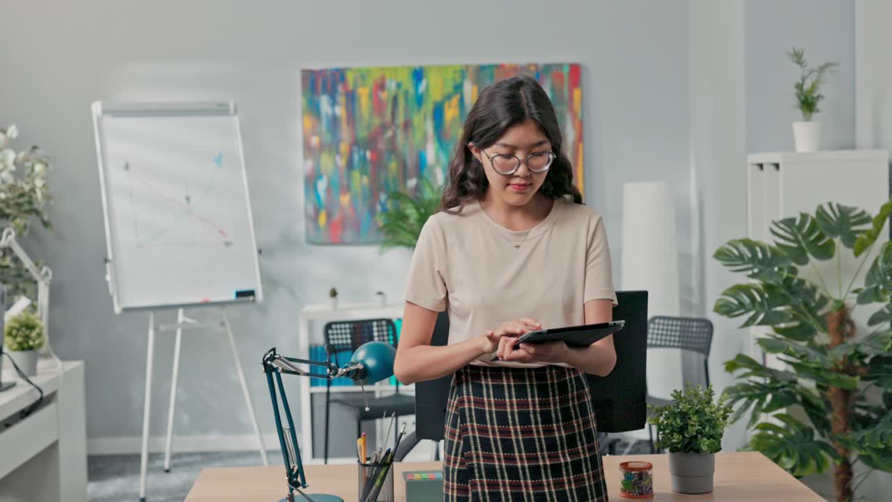Una bella donna con gli occhiali è in piedi davanti alla scrivania al centro dell'ufficio, controllando sul tablet gli ordini dell'azienda, il numero di ordini da completare e assegnando compiti ai colleghi.