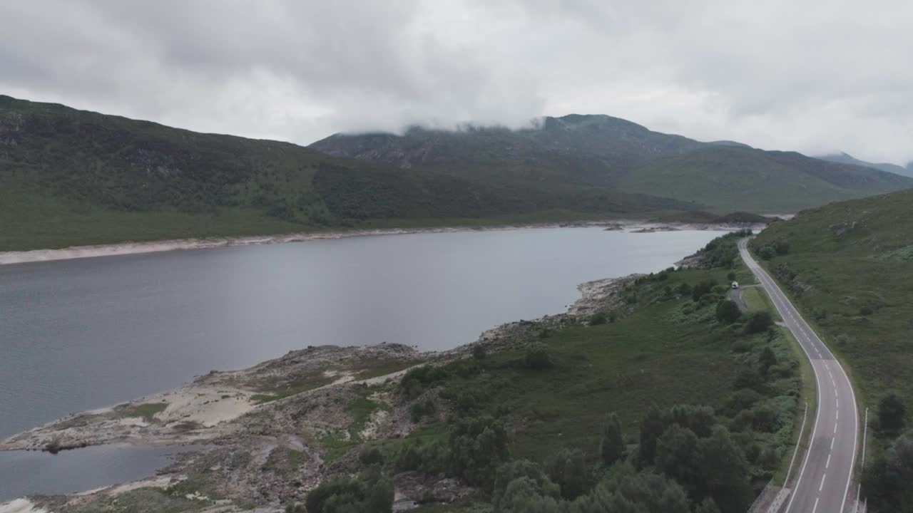 Wide drone shot with a campervan wild camping alongside Loch Beag, BBQ smoke with cloudy mountains in the background in Scottish Highlands