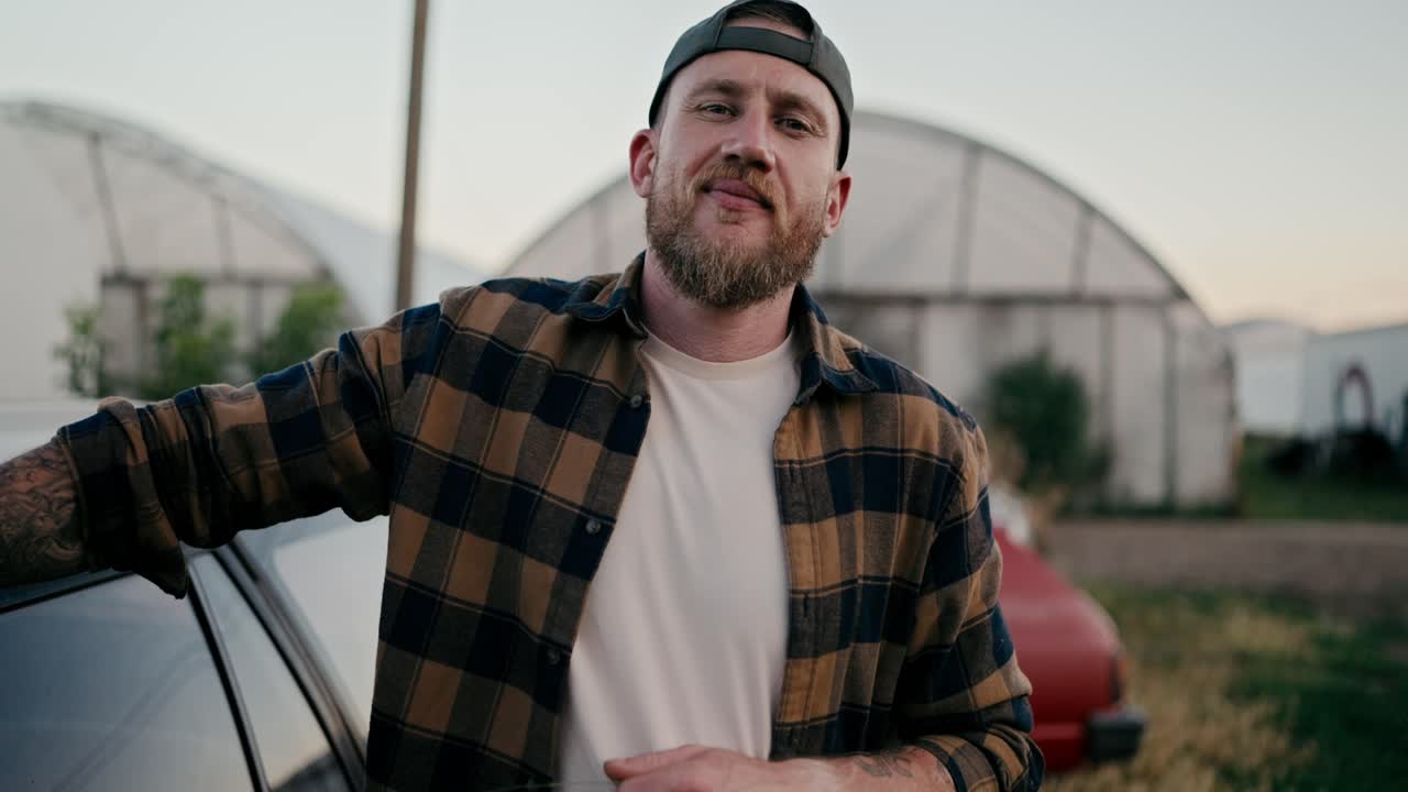 Portrait of a happy farmer guy in a cap and a plaid shirt posing near a car among greenhouses and fields on a farm
