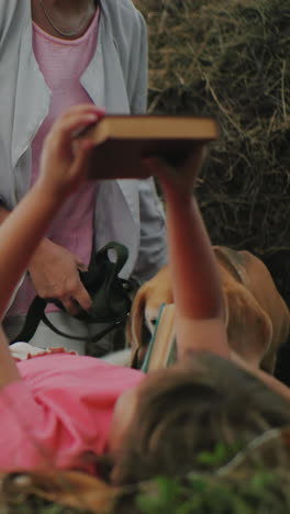 mujer de mediana edad entregando una novela a su hija tendida en el suelo mientras otro niño recoge libros, familia unida, disfrutando del tiempo en la naturaleza, rodeada de campos de cultivo abiertos y balas de heno