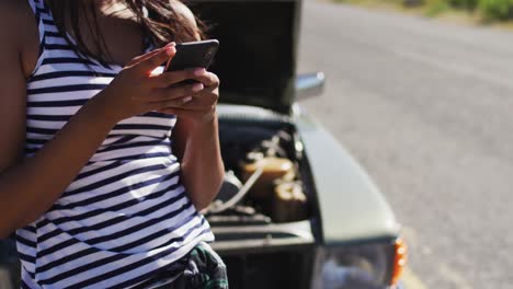 mujer afroamericana usando un teléfono inteligente mientras estaba de pie cerca de su coche averiado en la carretera