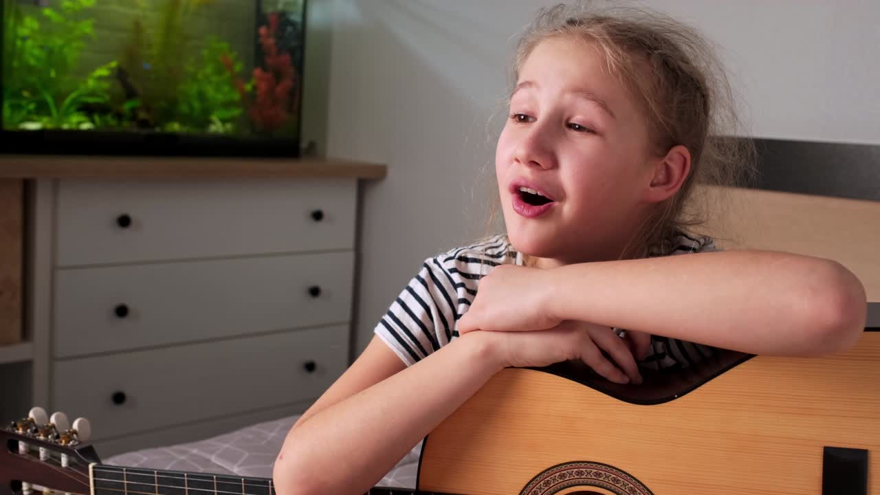 White teenage girl sings song leaning on acoustic guitar while sitting in room.