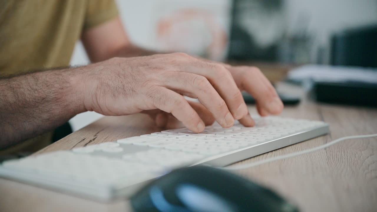 Male hands type on a slim computer keyboard at a wooden desk, with a mouse and monitor nearby conveying focused office work, typing, remote work and everyday productivity in a modern workspace. 4K